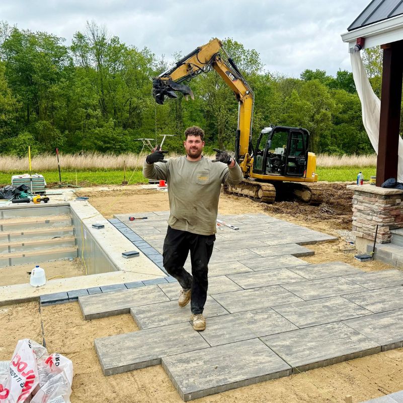 Construction worker posing on a new patio under construction near an excavator and a house.
