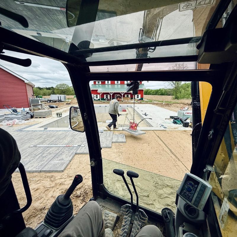 View from inside construction vehicle with a worker placing concrete slabs near a pool on sandy ground.