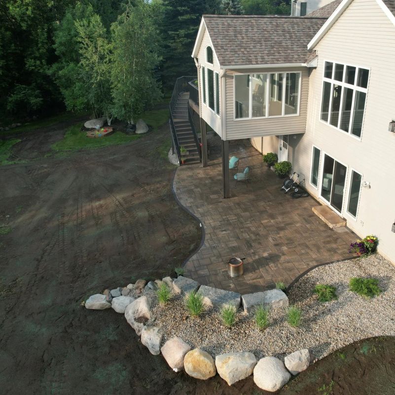 Patio with stone border and plants around a two-story house with large windows and outdoor seating.