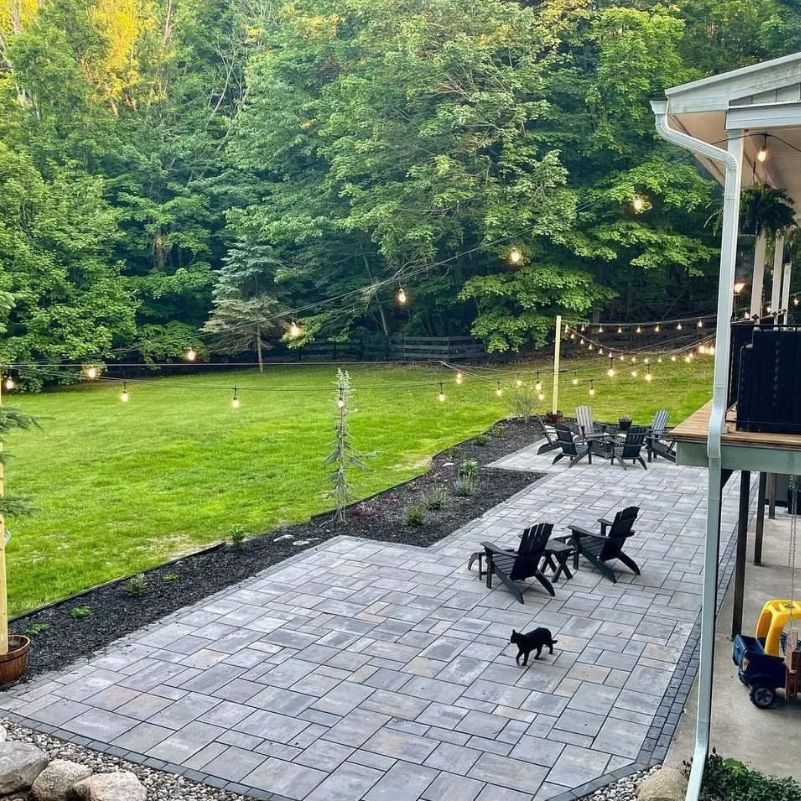 Backyard patio with black chairs, hanging string lights, green lawn, and a small black cat walking on stone tiles.