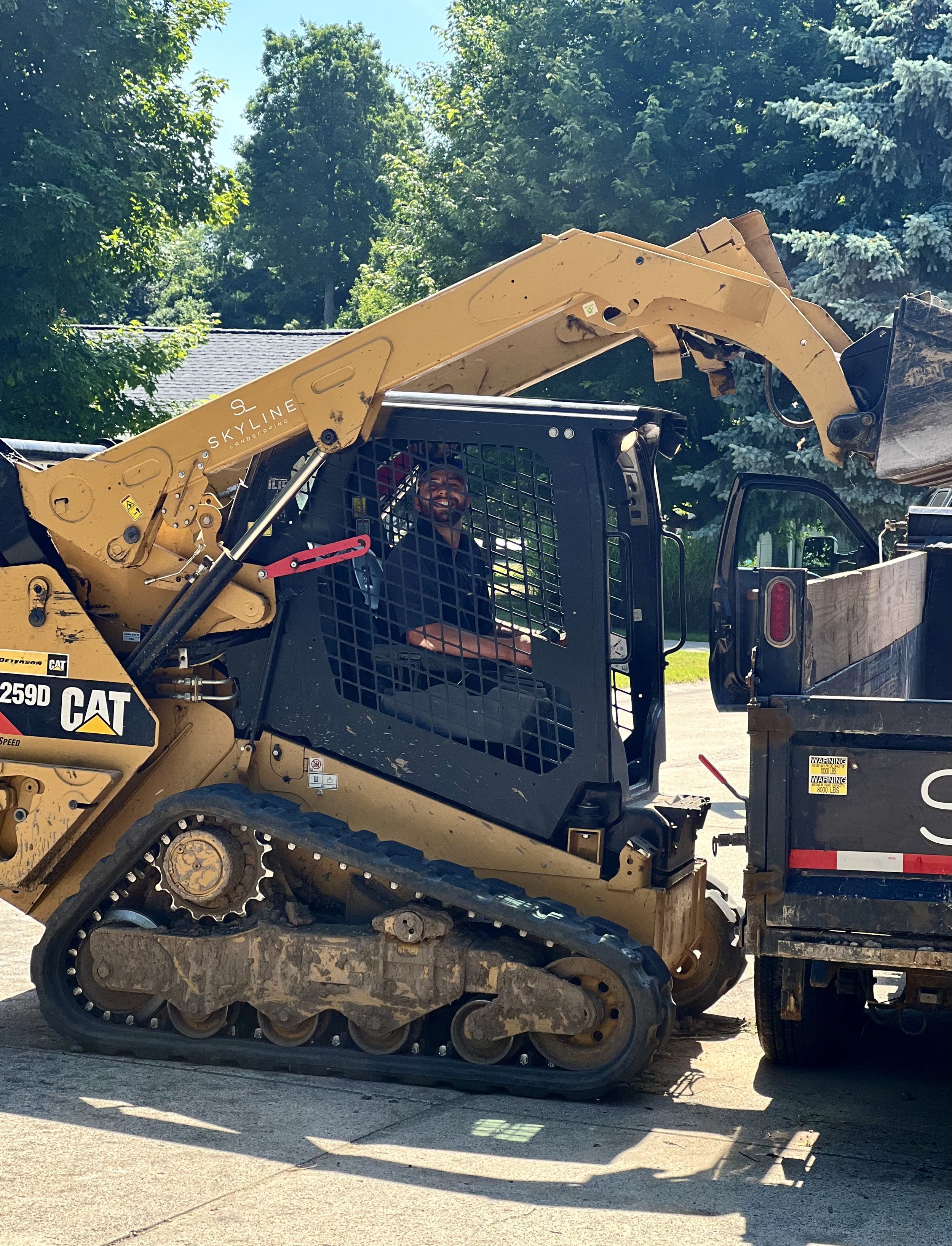 Man wearing a dark shirt sitting inside a CAT skid steer loader with a raised bucket on a sunny day.
