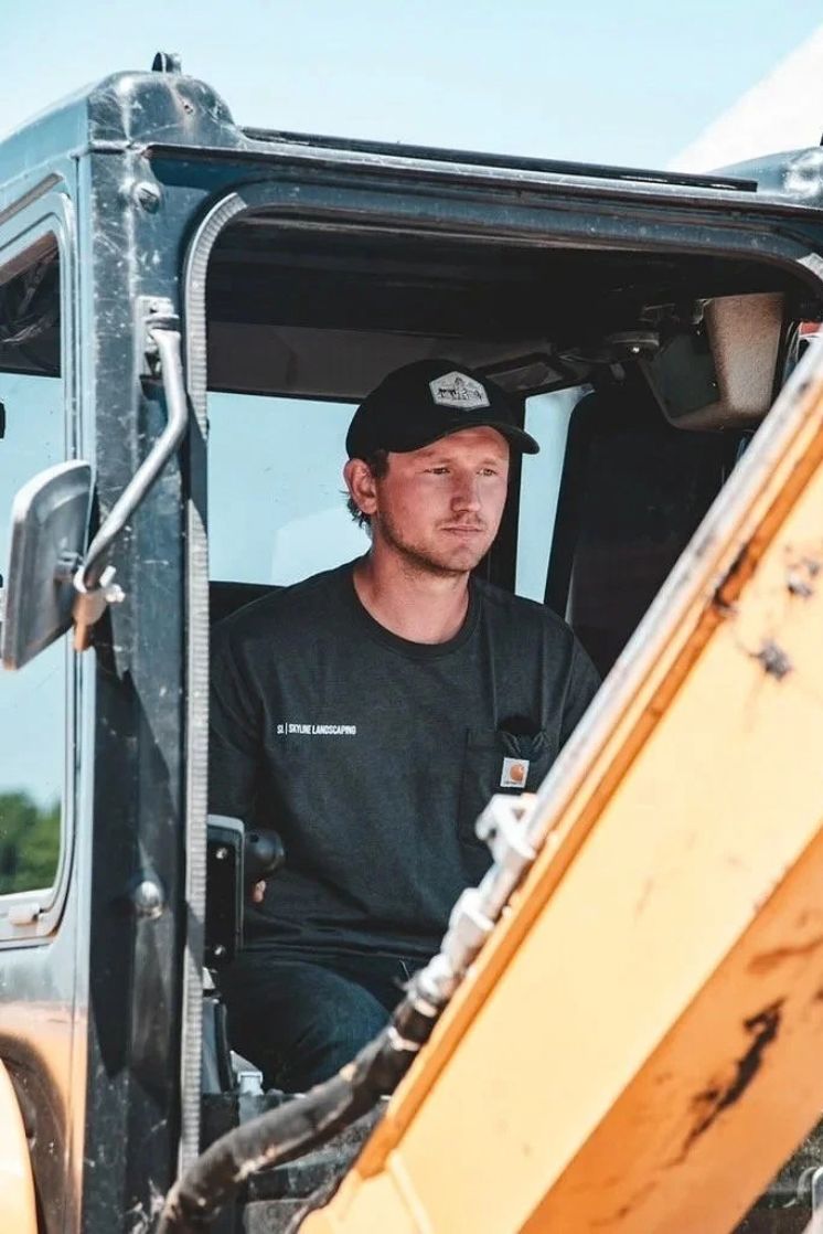 Man wearing a black cap and shirt operating heavy construction equipment inside a machinery cabin.
