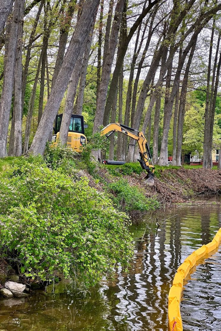 Yellow excavator among tall trees working near a calm water body with a yellow barrier in the water.