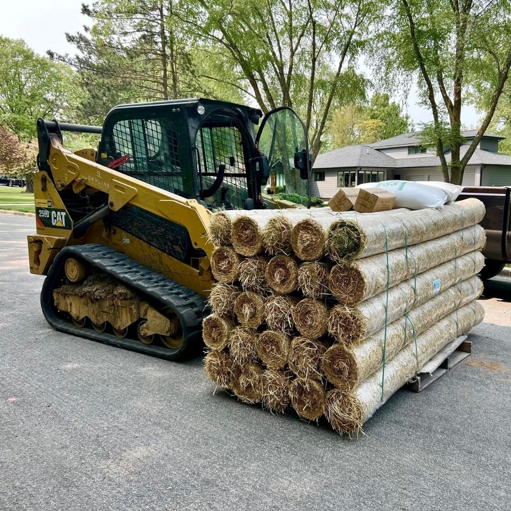 Tracked Caterpillar compact loader parked near a pallet of rolled straw erosion control blankets.