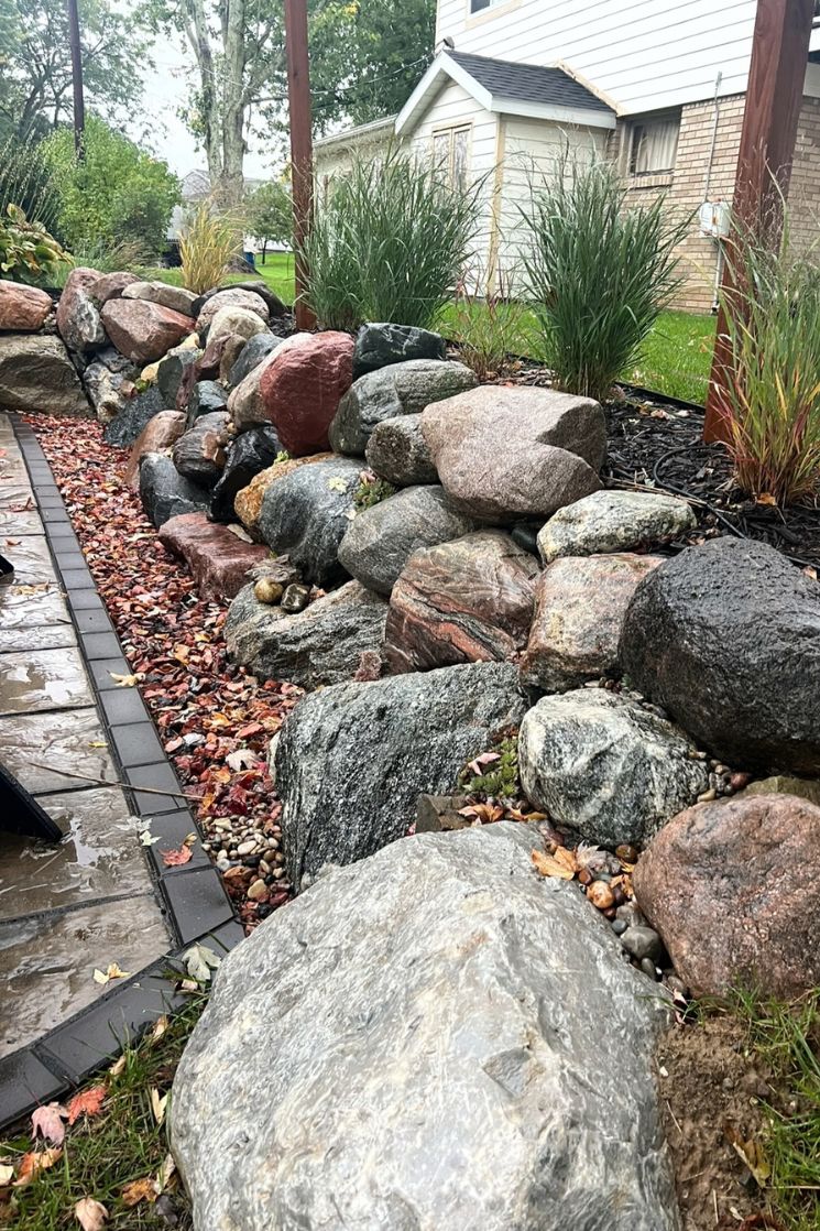 Rock garden with large boulders, ornamental grasses, and a wet stone tile walkway beside a house.
