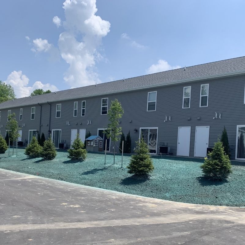 Row of gray townhouses with small trees and blue-green ground cover in front under a partly cloudy sky.