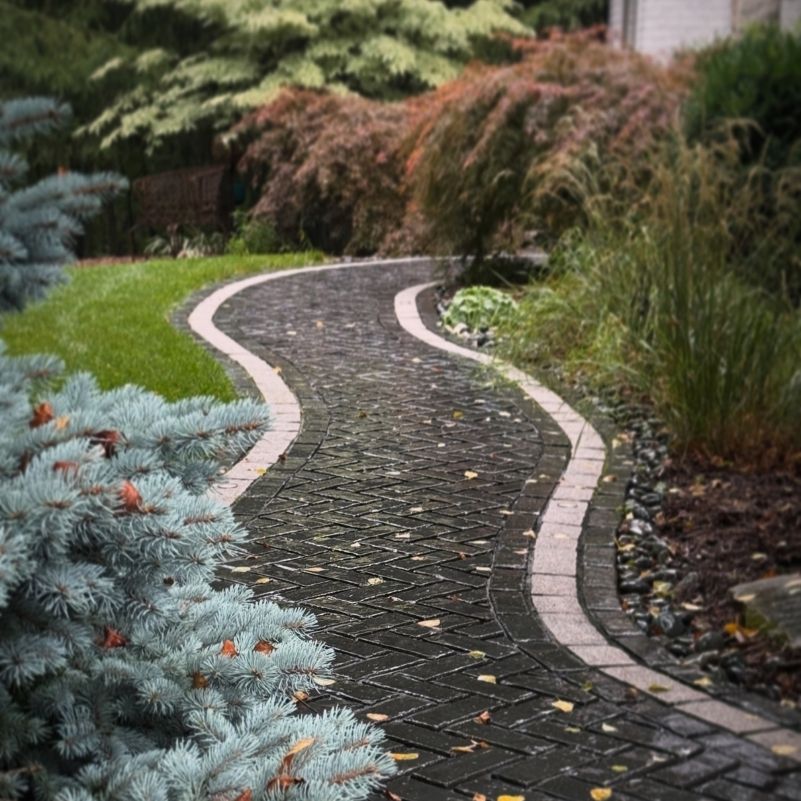Curved paved pathway with bricks bordered by green grass, bushes, and plants on both sides.