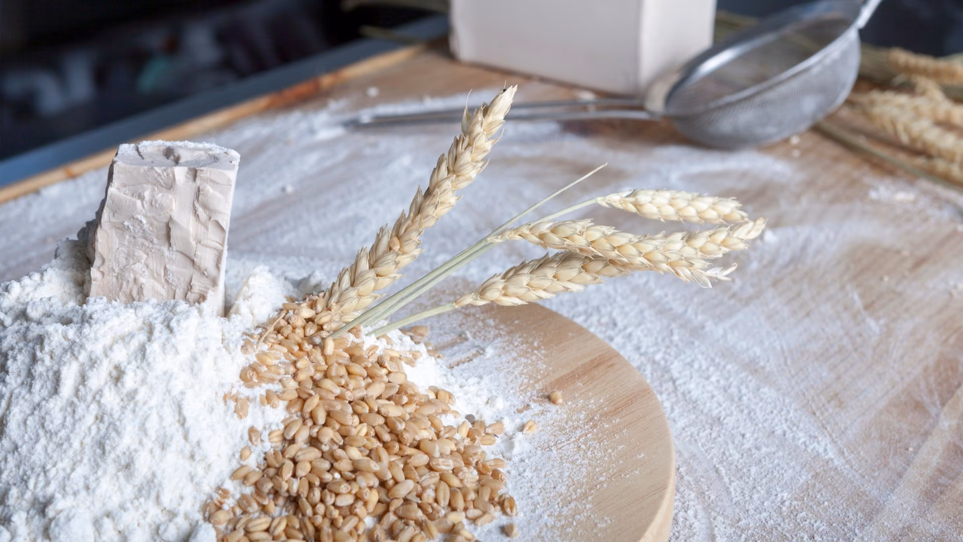 Wheat grains and wheat stalks on flour-covered wooden surface with baking ingredients and sieve in the background.