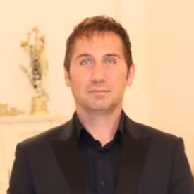 Portrait of a man with short brown hair wearing a black suit jacket and shirt against a cream-colored background.