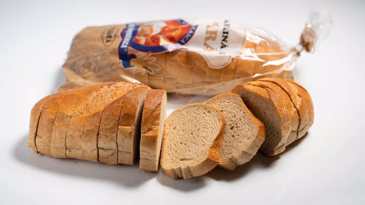 Sliced loaf of brown bread on a white surface with its plastic packaging in the background.