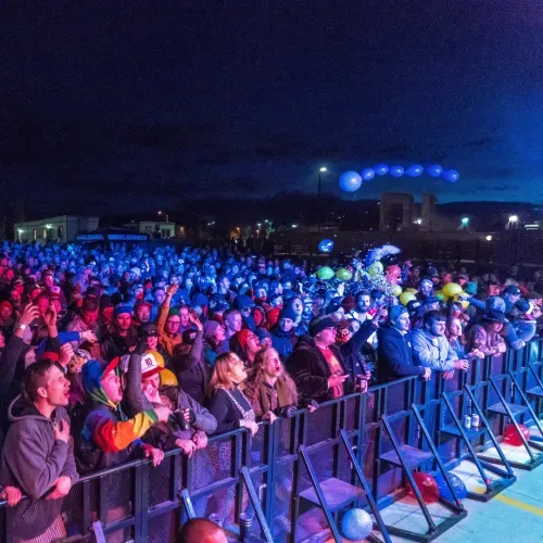 A large crowd at an outdoor night concert, pressed against a barrier under blue and red stage lighting, with balloons floating above the audience.