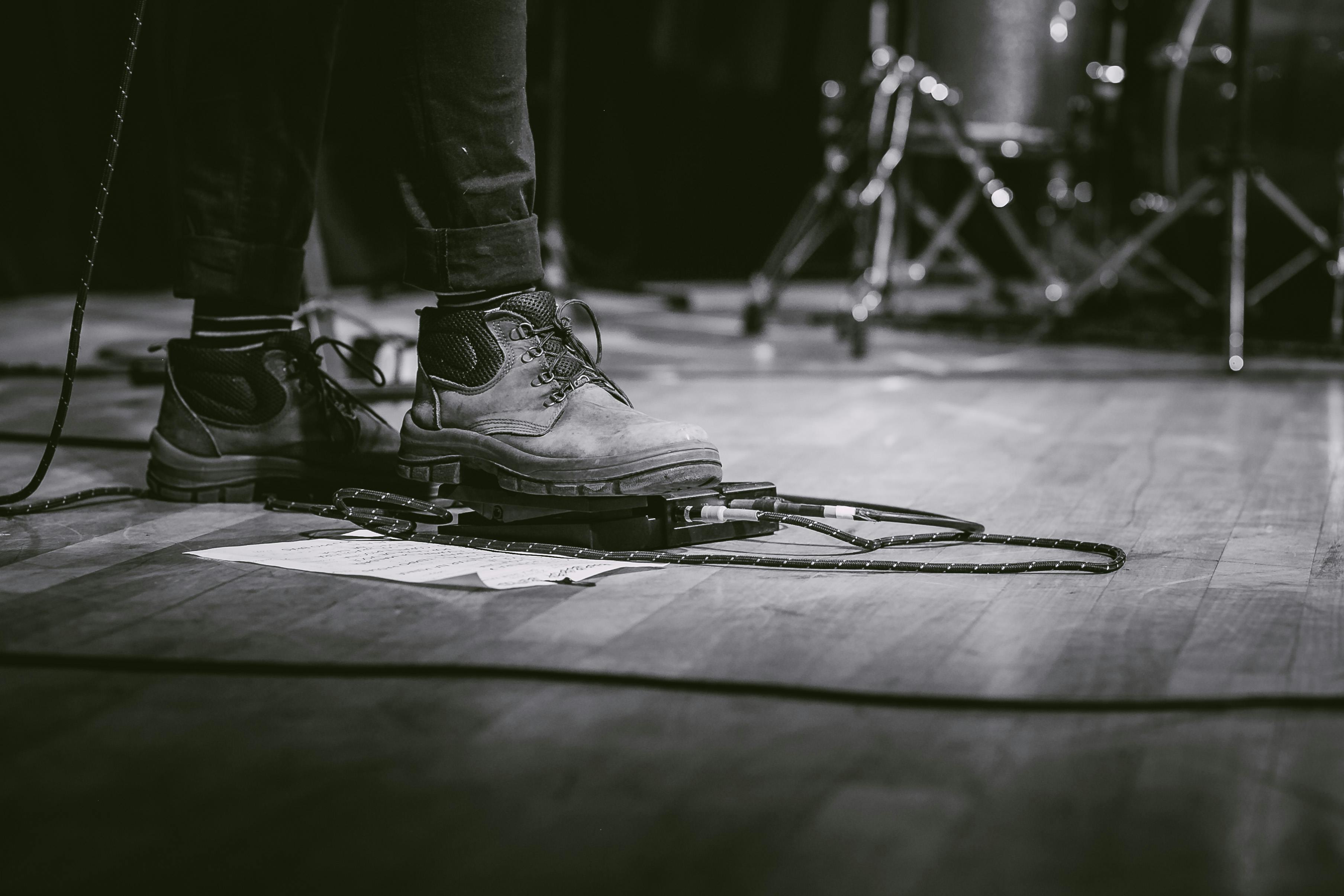 Close-up of a musician’s boots operating a guitar effects pedal on a wooden stage floor, with cables and a handwritten setlist nearby — capturing the technical setup of a live performance.