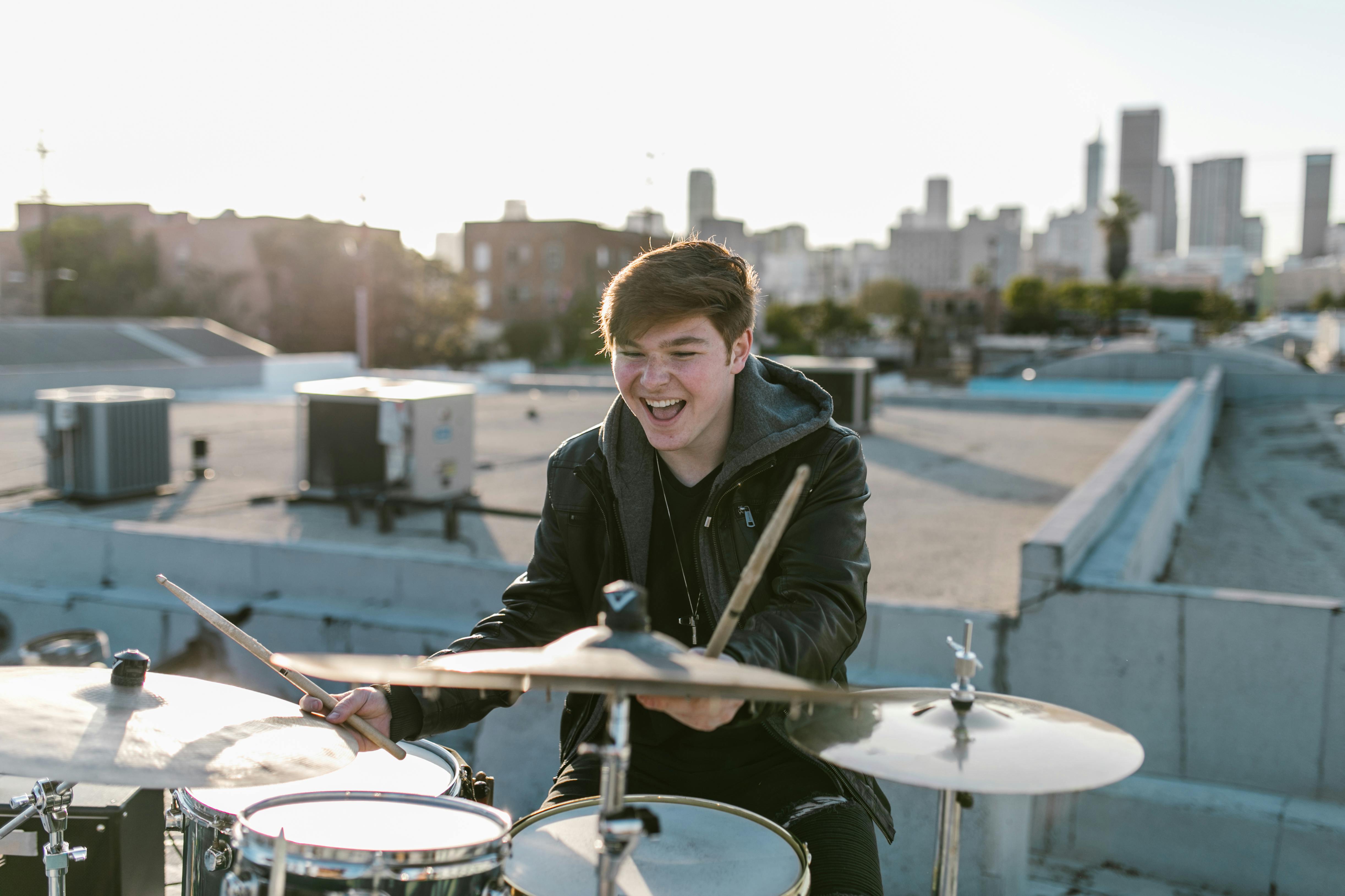 Young man playing a drum set on a rooftop in an urban setting, wearing a black leather jacket, with city skyline and HVAC units in the background.