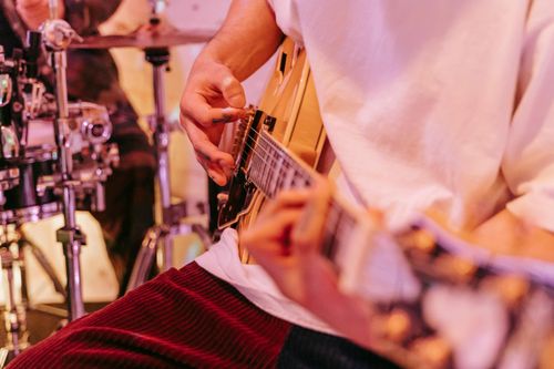 Person playing an electric guitar in a live band setting, wearing a white shirt and red corduroy pants, with a drum set visible in the background.