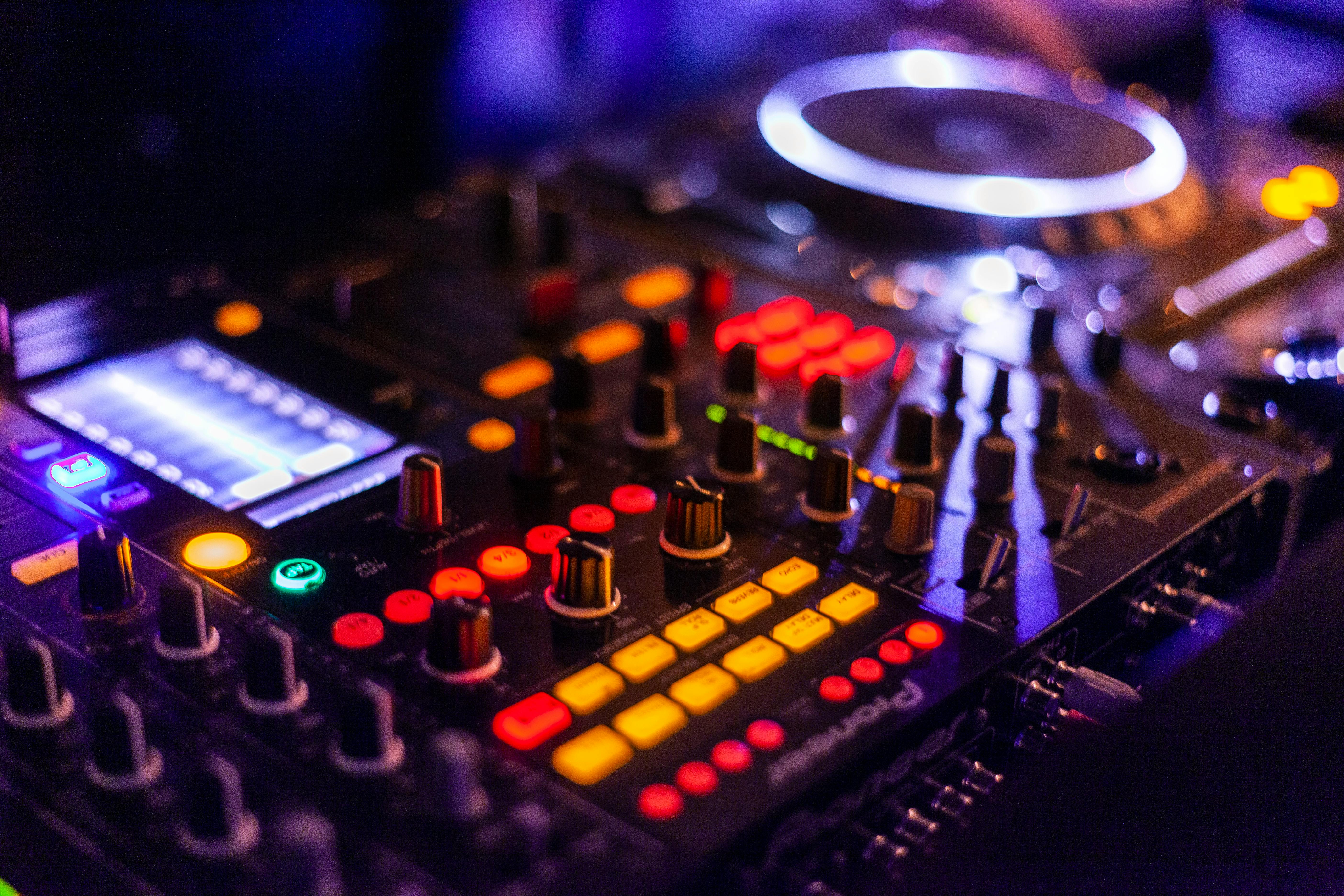 Close-up of a Pioneer DJ mixing console with colorful LED lights, knobs, buttons, and a glowing jog wheel, used for live music mixing and audio effects.