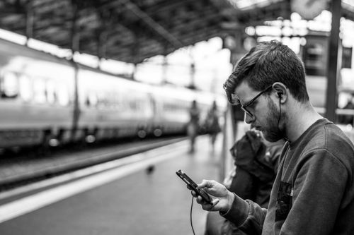 Black-and-white photo of a bearded man with glasses and earphones sitting on a train station bench, looking at his smartphone. A train is visible in the background with blurred commuters and overhead roof structure, capturing a moment of modern urban trave