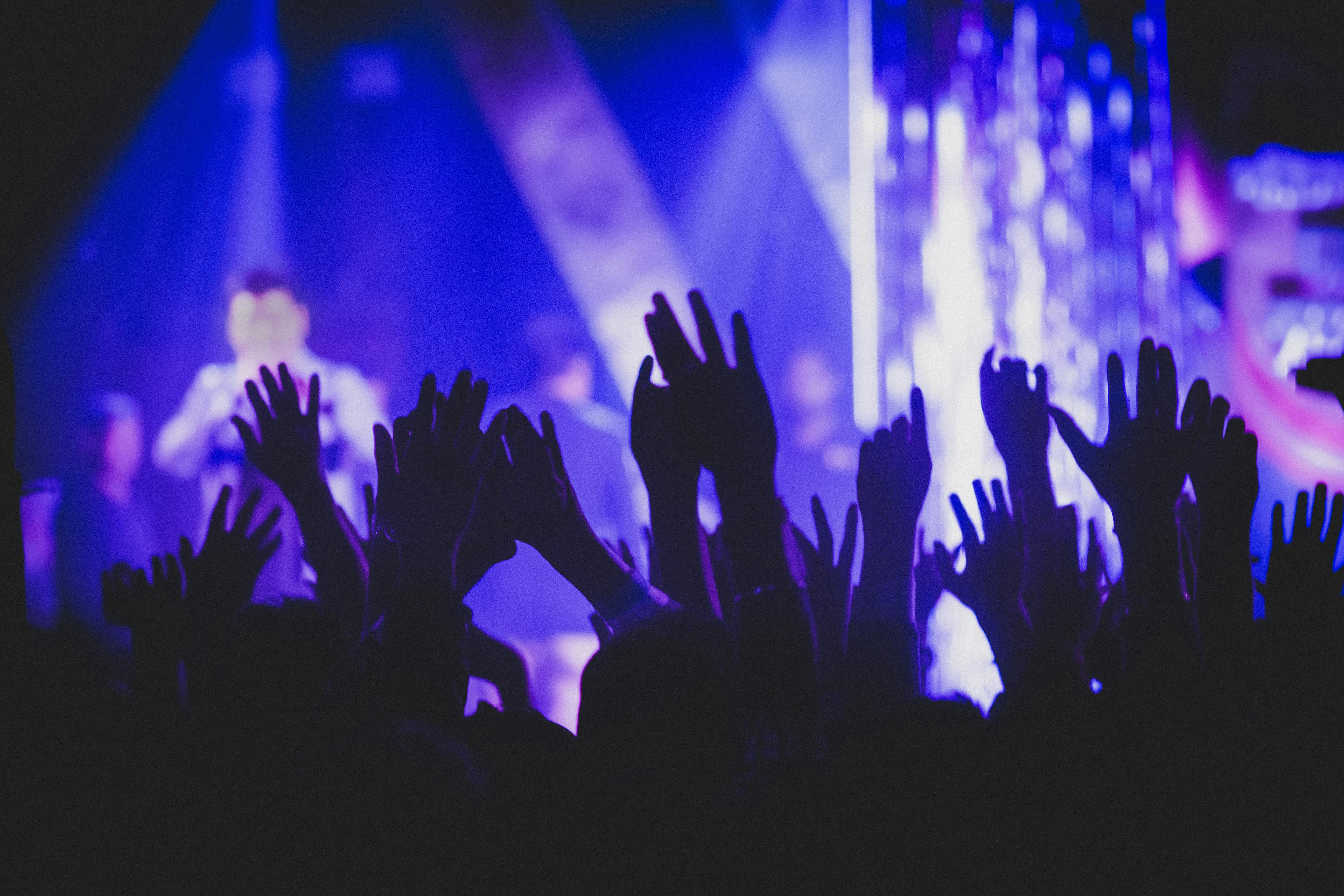 Energetic crowd at a live music concert with raised hands silhouetted in the foreground and performers illuminated by vibrant blue and purple stage lighting in the background.