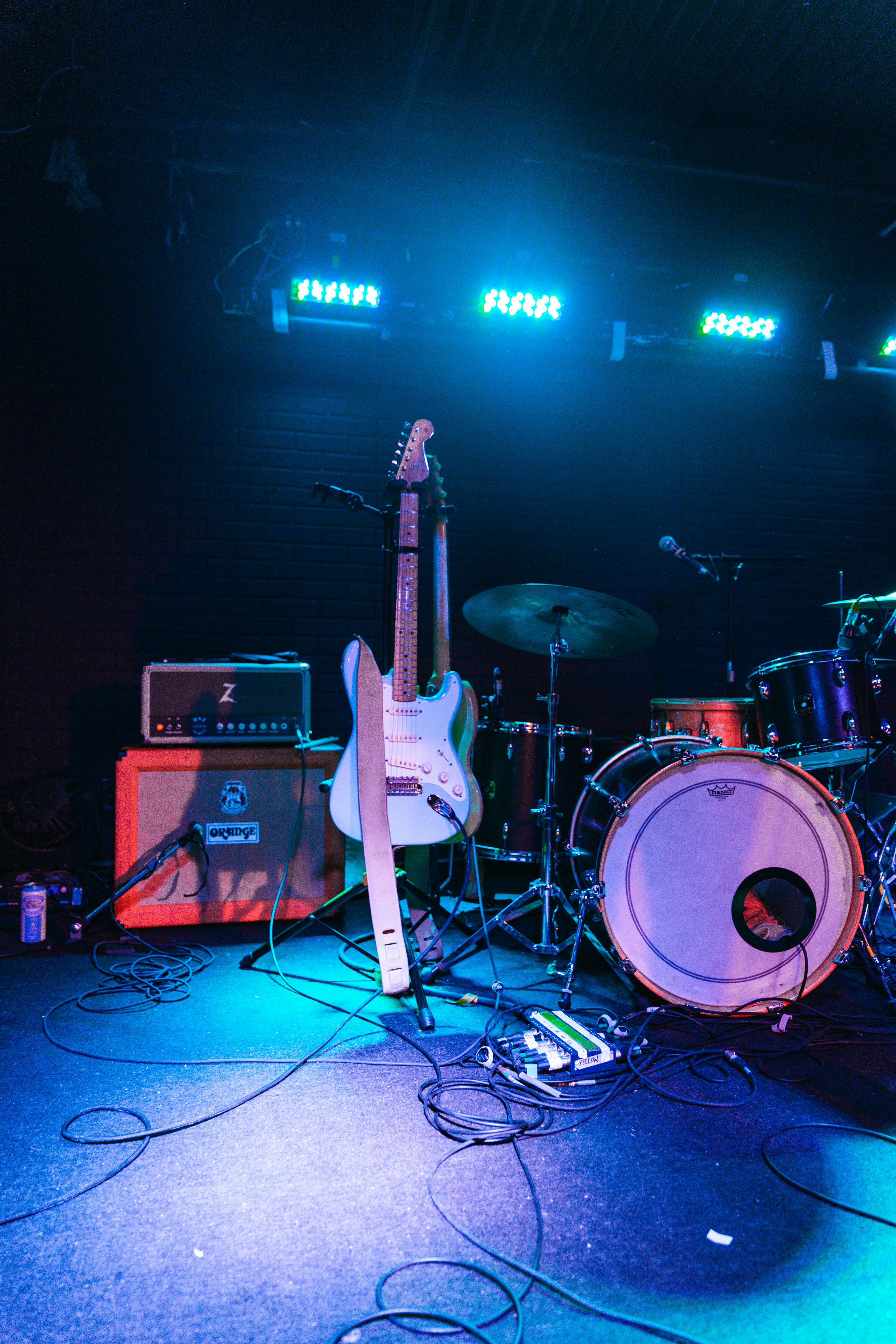 Stage setup for a live music performance featuring a white electric guitar on a stand, a full drum kit, amplifier stack, pedalboard, and vibrant blue-green stage lighting.
