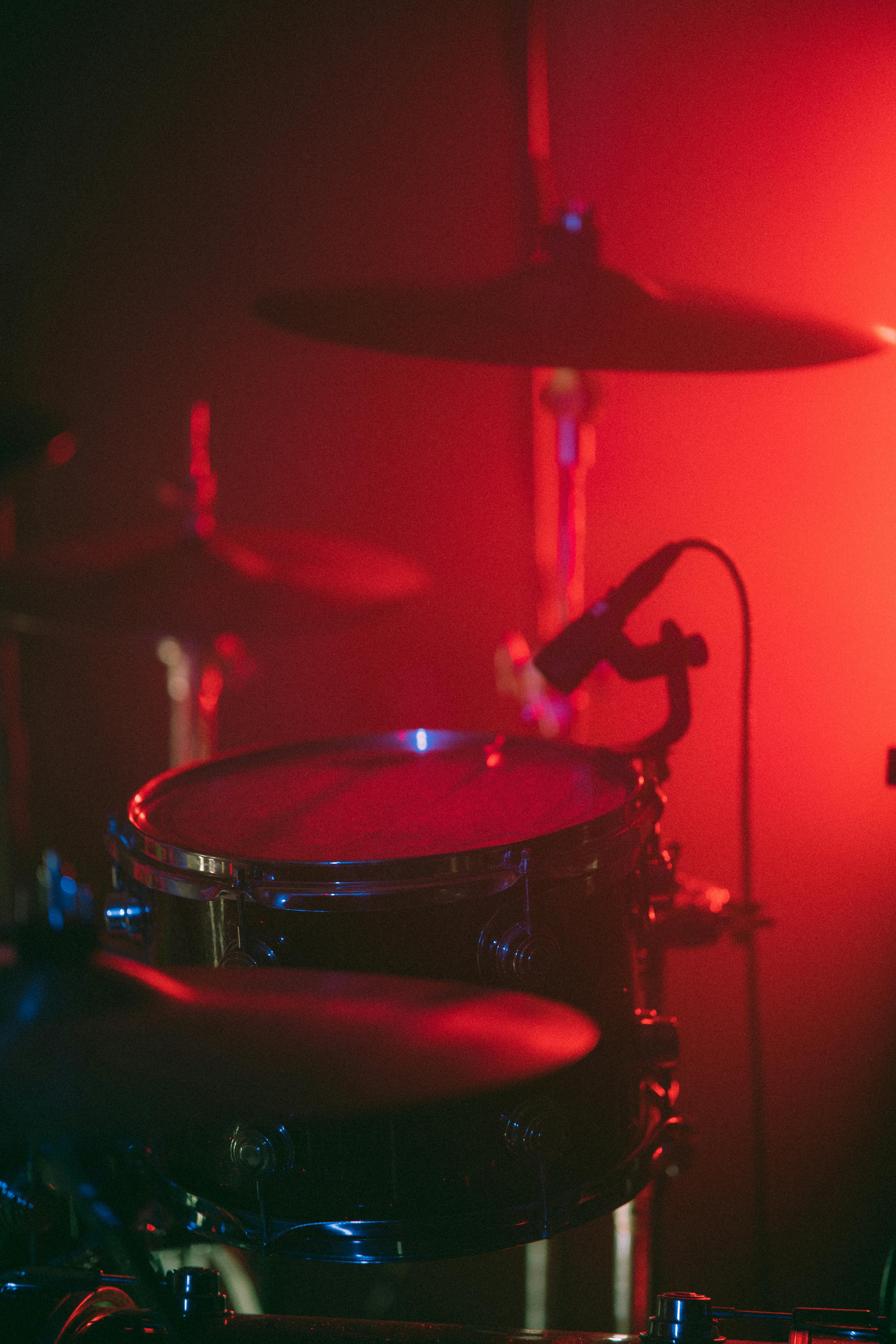 Close-up of a drum set under dramatic red and blue stage lighting, featuring snare, cymbal, and microphone in a live performance setting.