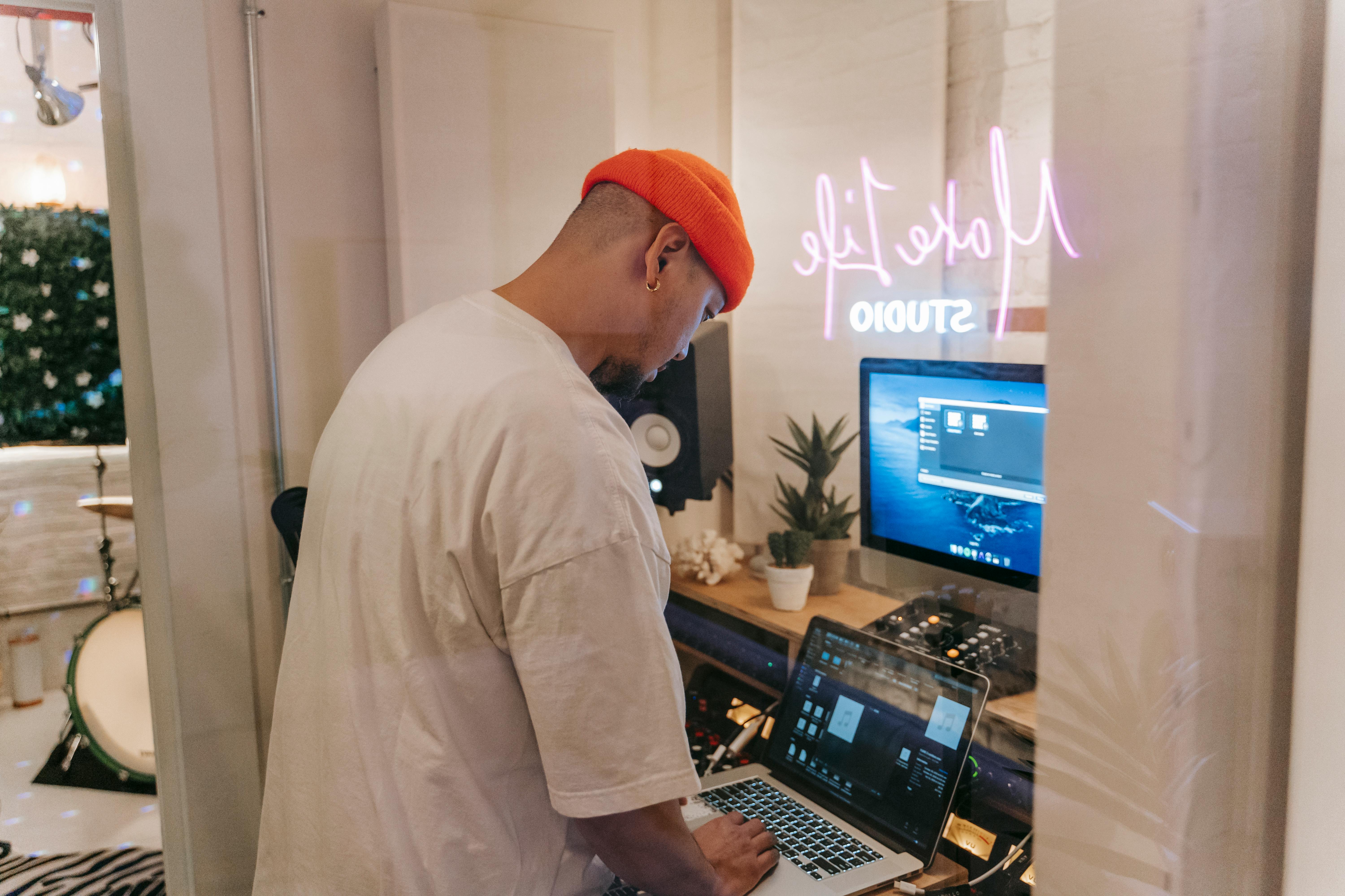 Person in red beanie working on music production in a modern studio with laptop, speakers, and neon sign.