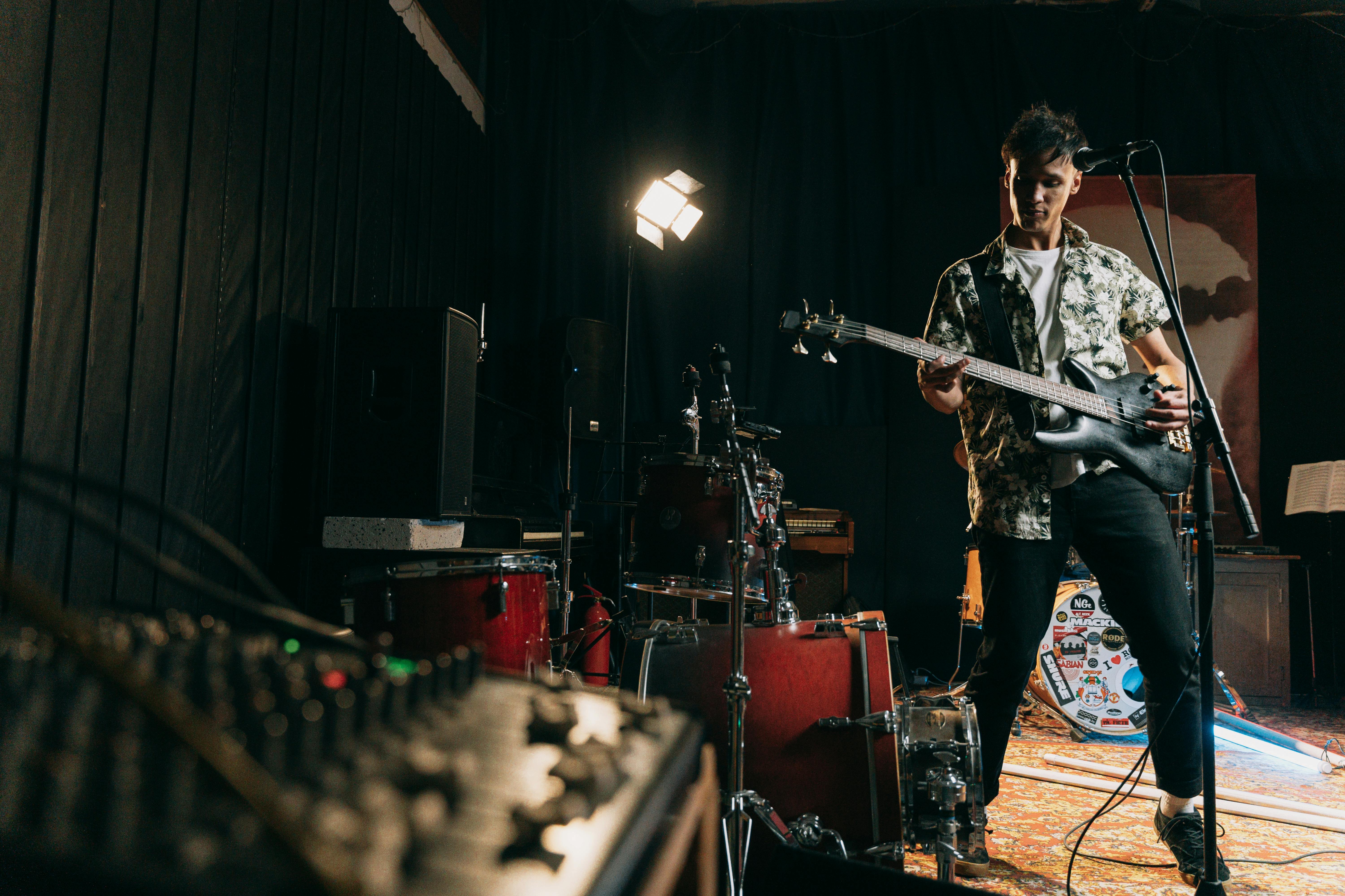 A musician plays an electric bass guitar on a small indoor stage, surrounded by drums, speakers, and audio equipment. Stage lights illuminate the scene as he performs in a casual, intimate rehearsal or live setup.