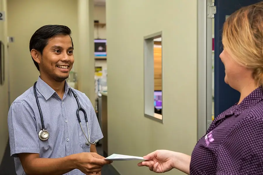 Male doctor giving papers to other staff member. They are both smiling with patient rooms in background.