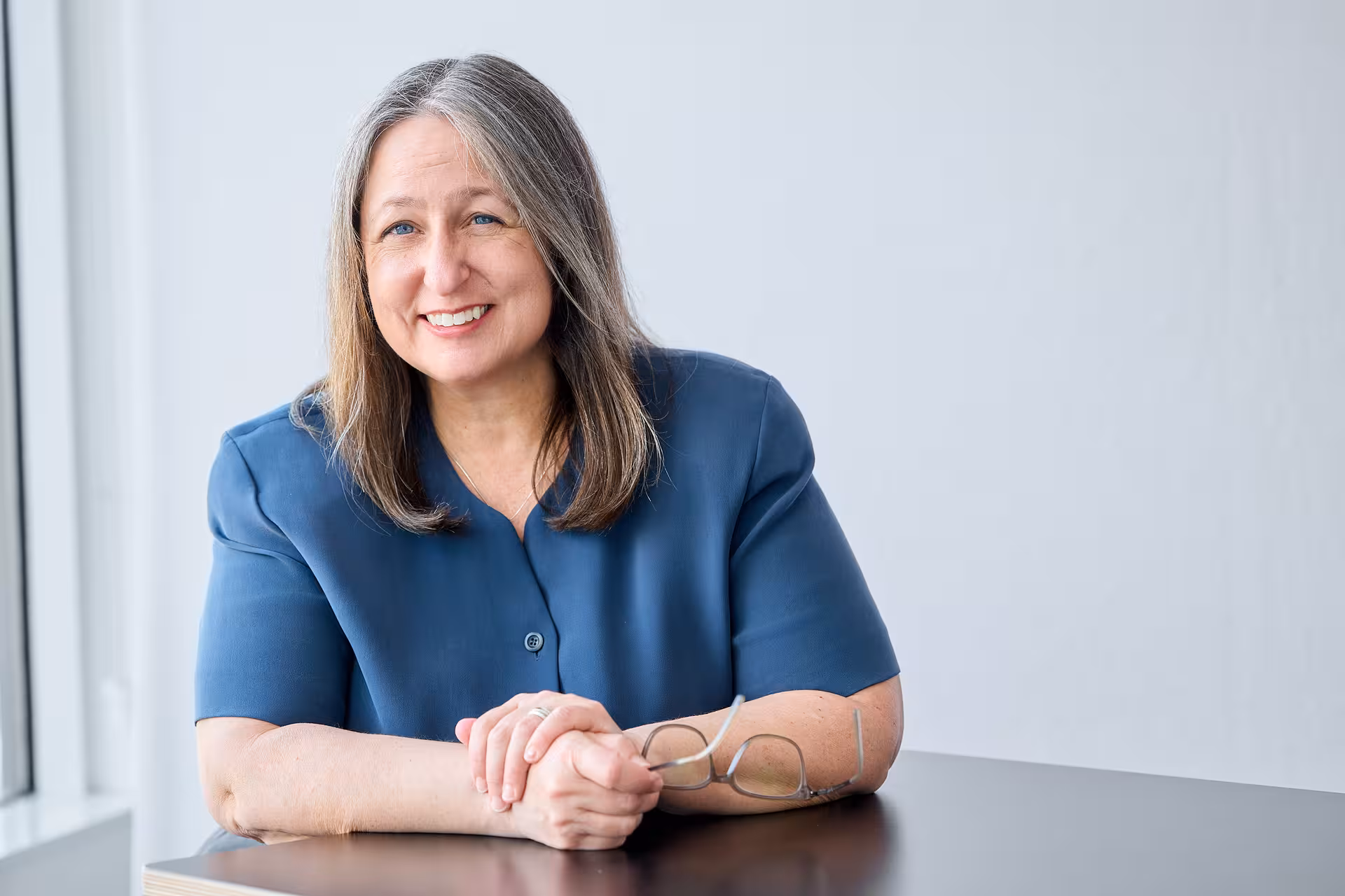 Portrait of woman with her hands folded on a table and holding glasses in one hand.