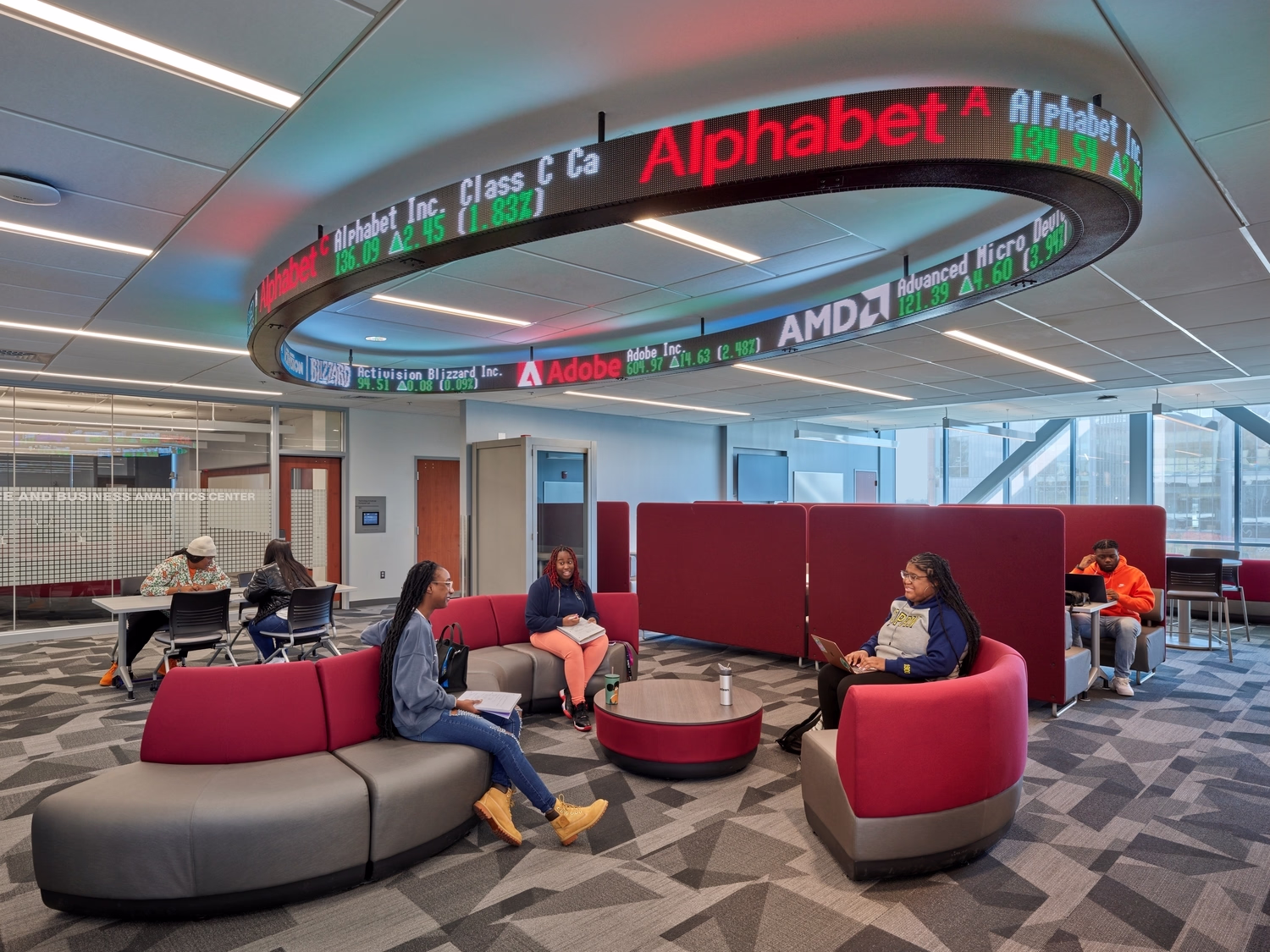 Below a three-dimensional circular Nasdaq ticker, students interact with each other inside a lounge.