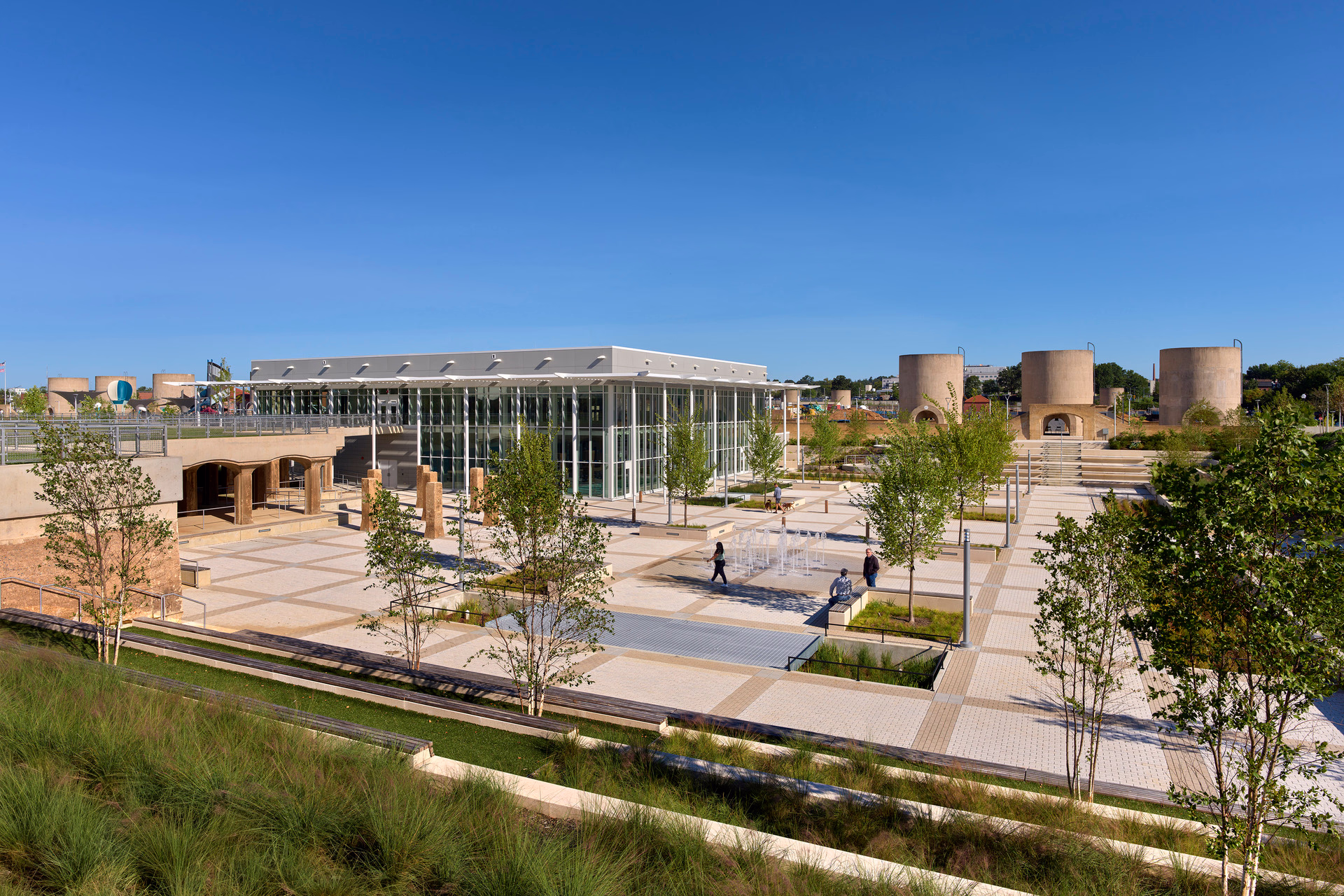 A photo of a plaza with a splash pad in front of a recreation center and historic infrastructure.
