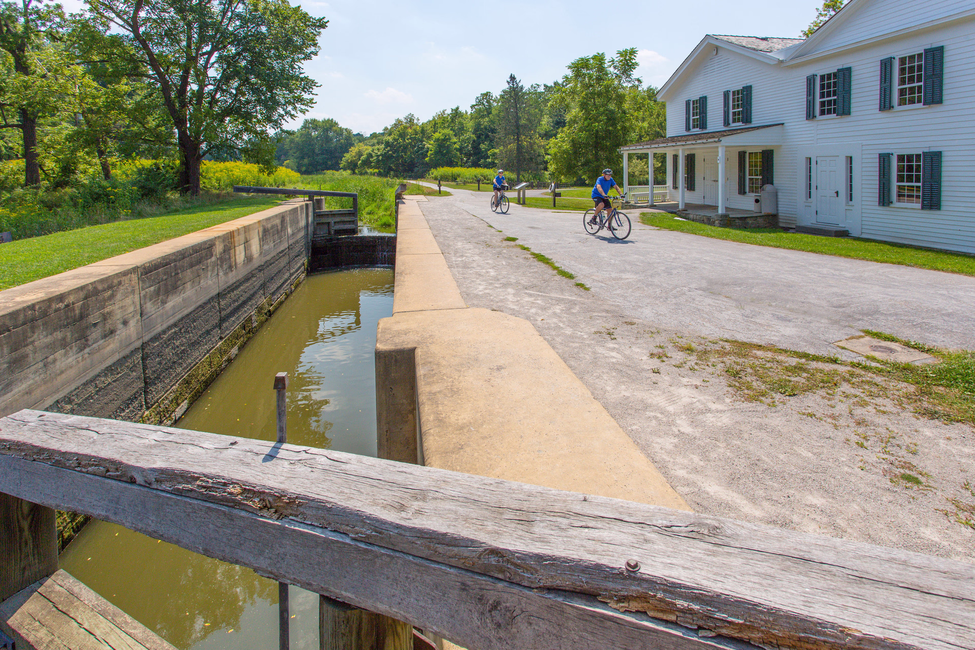 A photo of two people riding bicycles on a gravel path between a canal lock and a historic building.