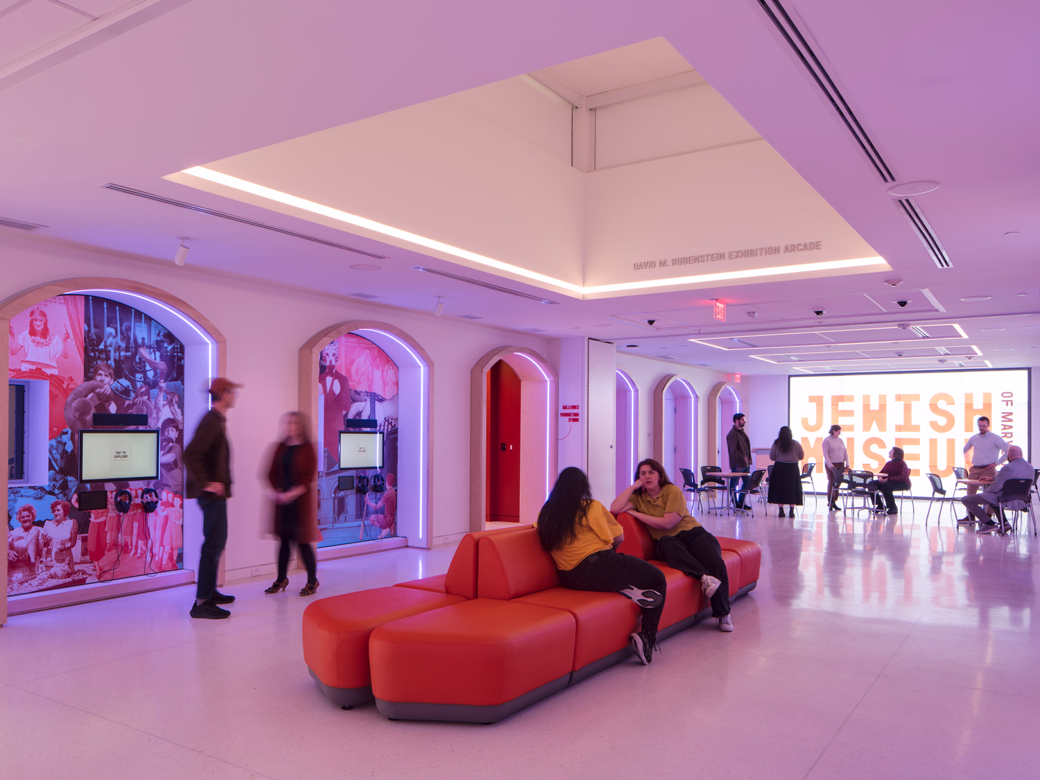 An interior photo of a museum atrium with people sitting on a couch, looking at exhibits, and seated at tables in front of a wall-size LCD screen.