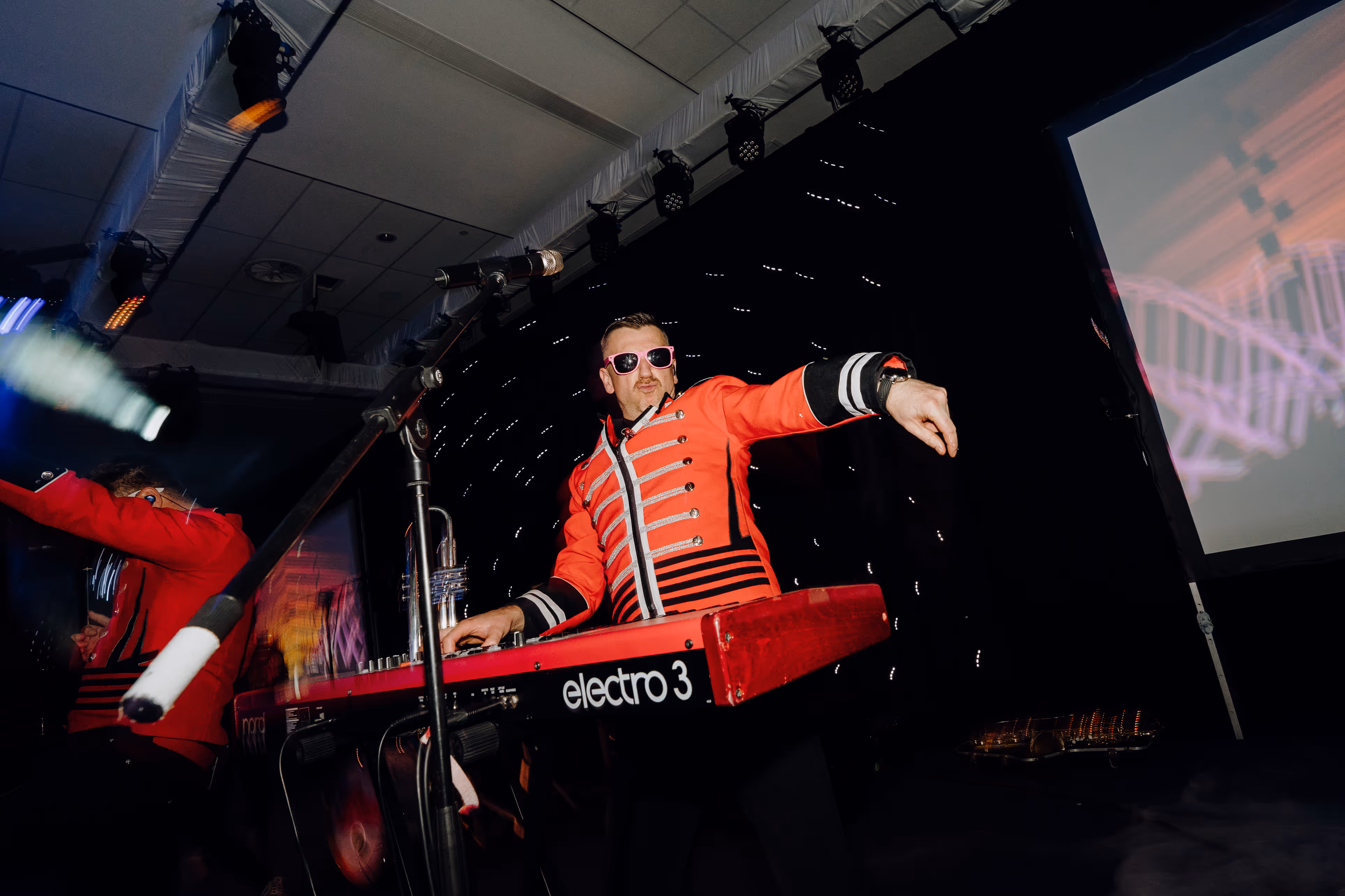Soniq Empires' keyboardist in a red jacket playing a Nord Electro keyboard and engaging the crowd during a lively awards event performance.