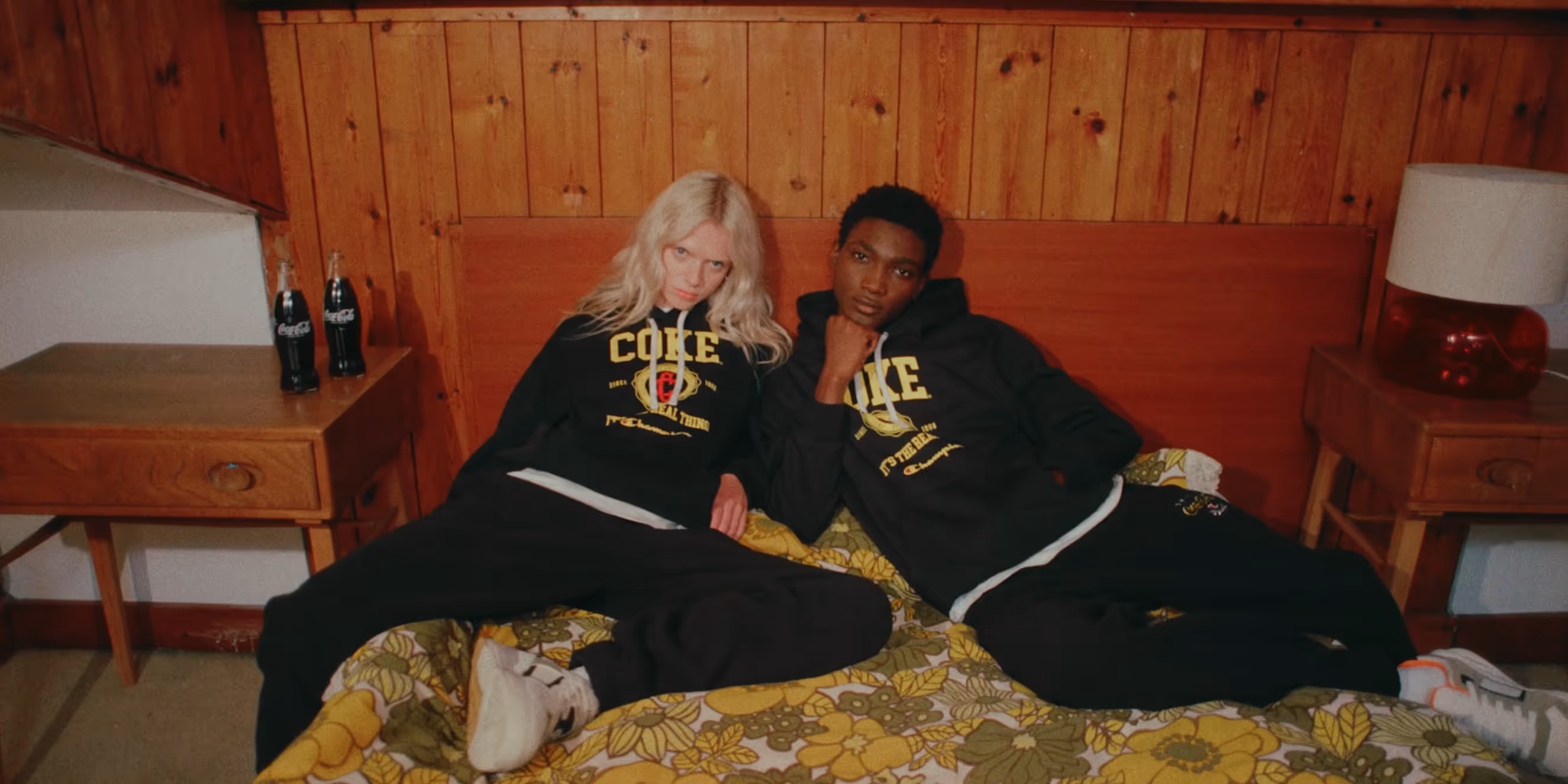 Two people wearing black hoodies with yellow Coca-Cola and Champion logos reclining on a bed with a floral blanket in a wood-paneled room.