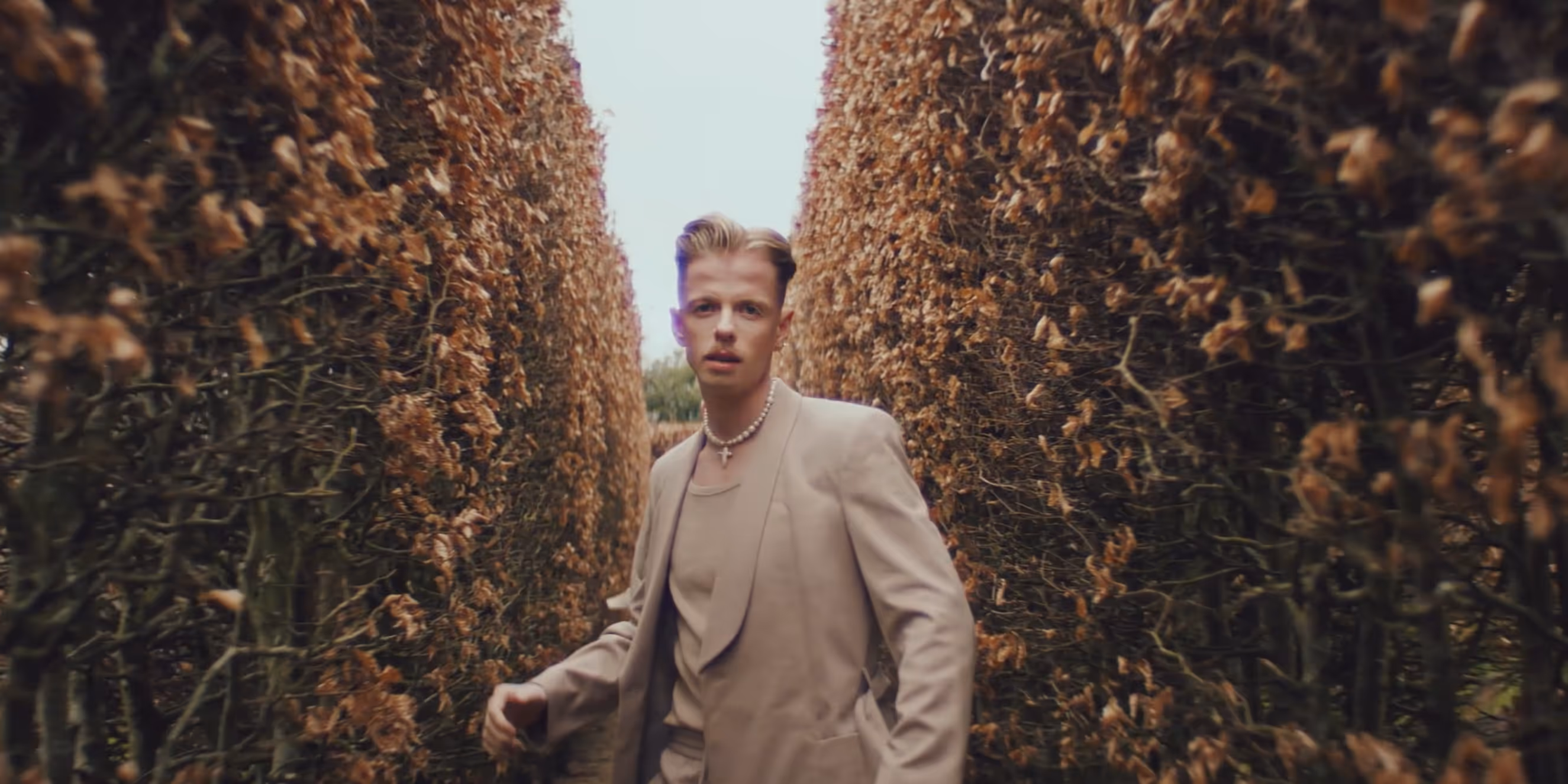 Young man in a beige suit and pearl necklace standing between two tall rows of dried brown bushes.