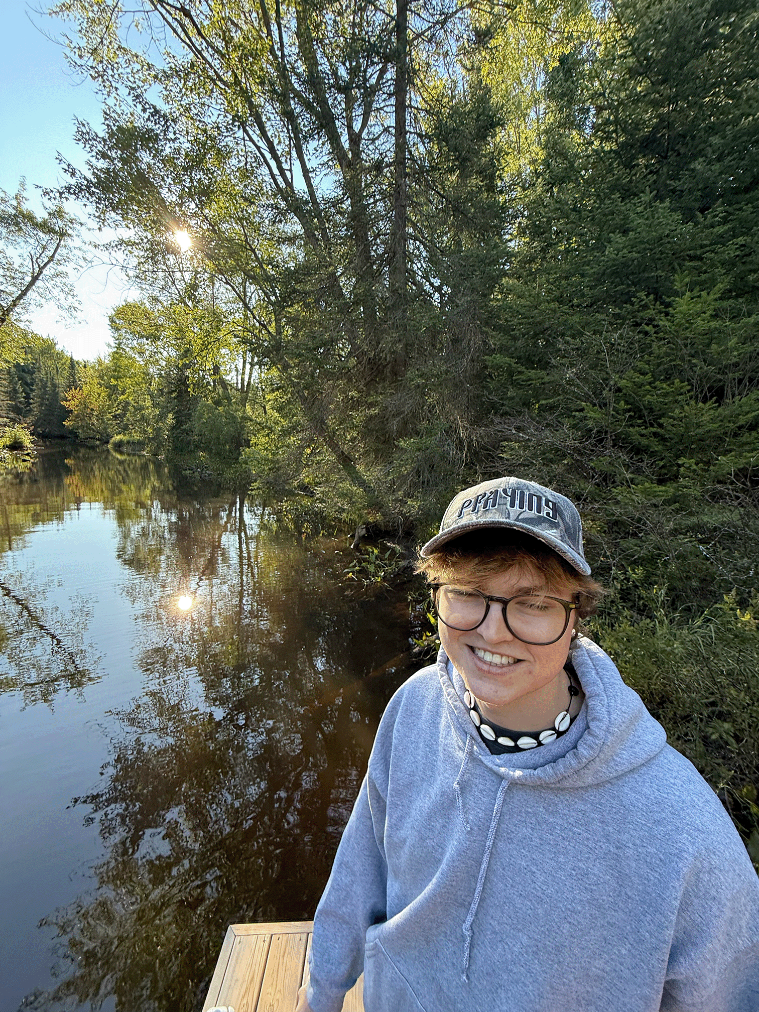 Person wearing glasses, a gray hoodie, a shell necklace, and a camo cap standing on a wooden dock by a forested water body.