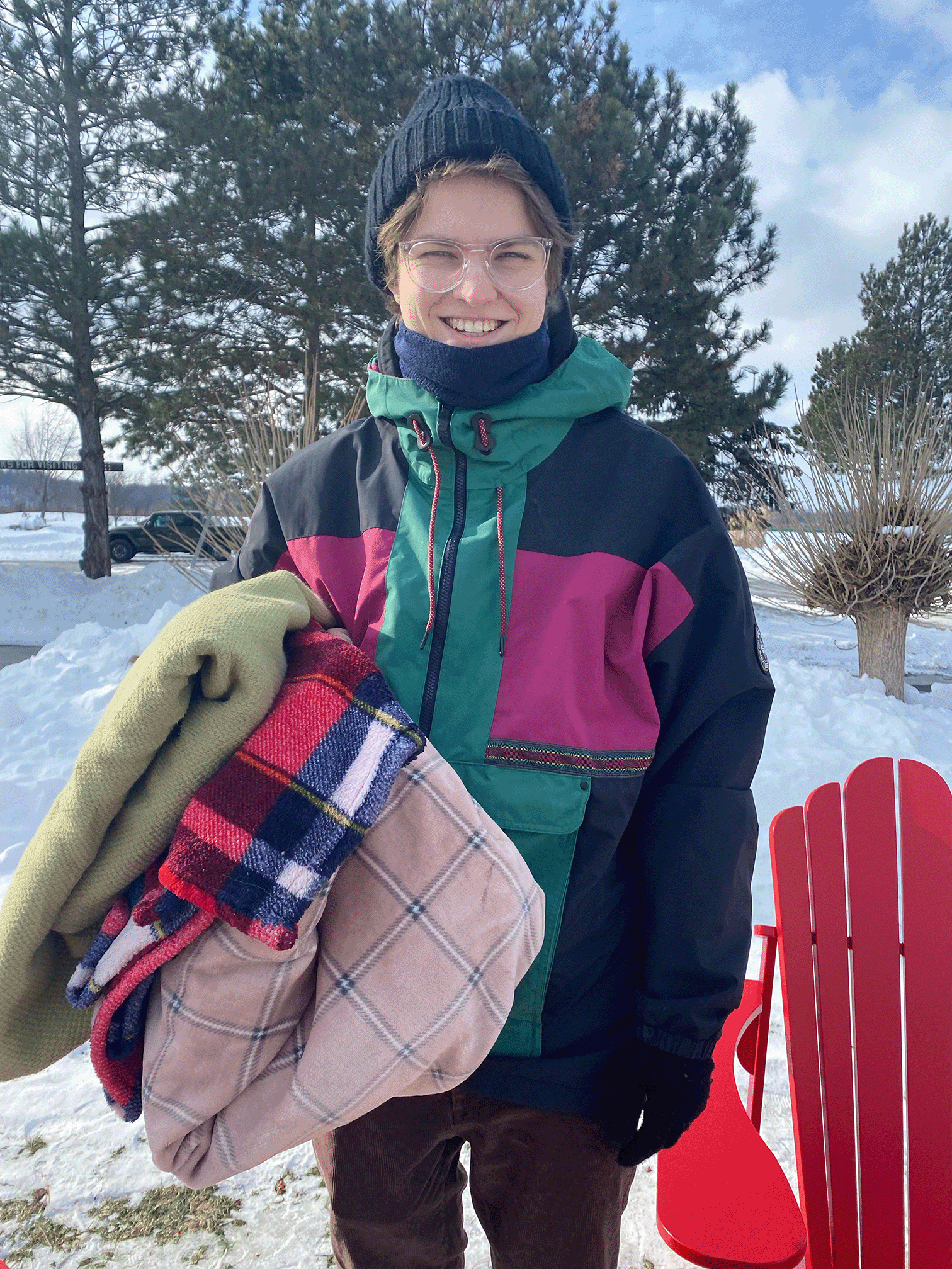 Smiling person wearing glasses, a black beanie, and a colorful winter jacket holding folded blankets outside in snowy weather.