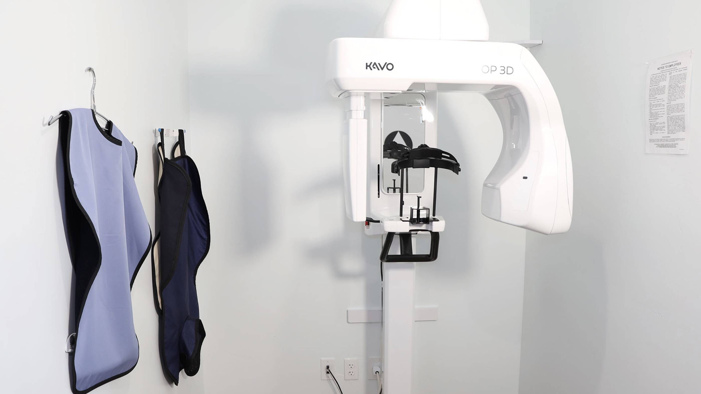 KaVo OP 3D dental X-ray machine in a white room with two protective lead aprons hanging on the wall.