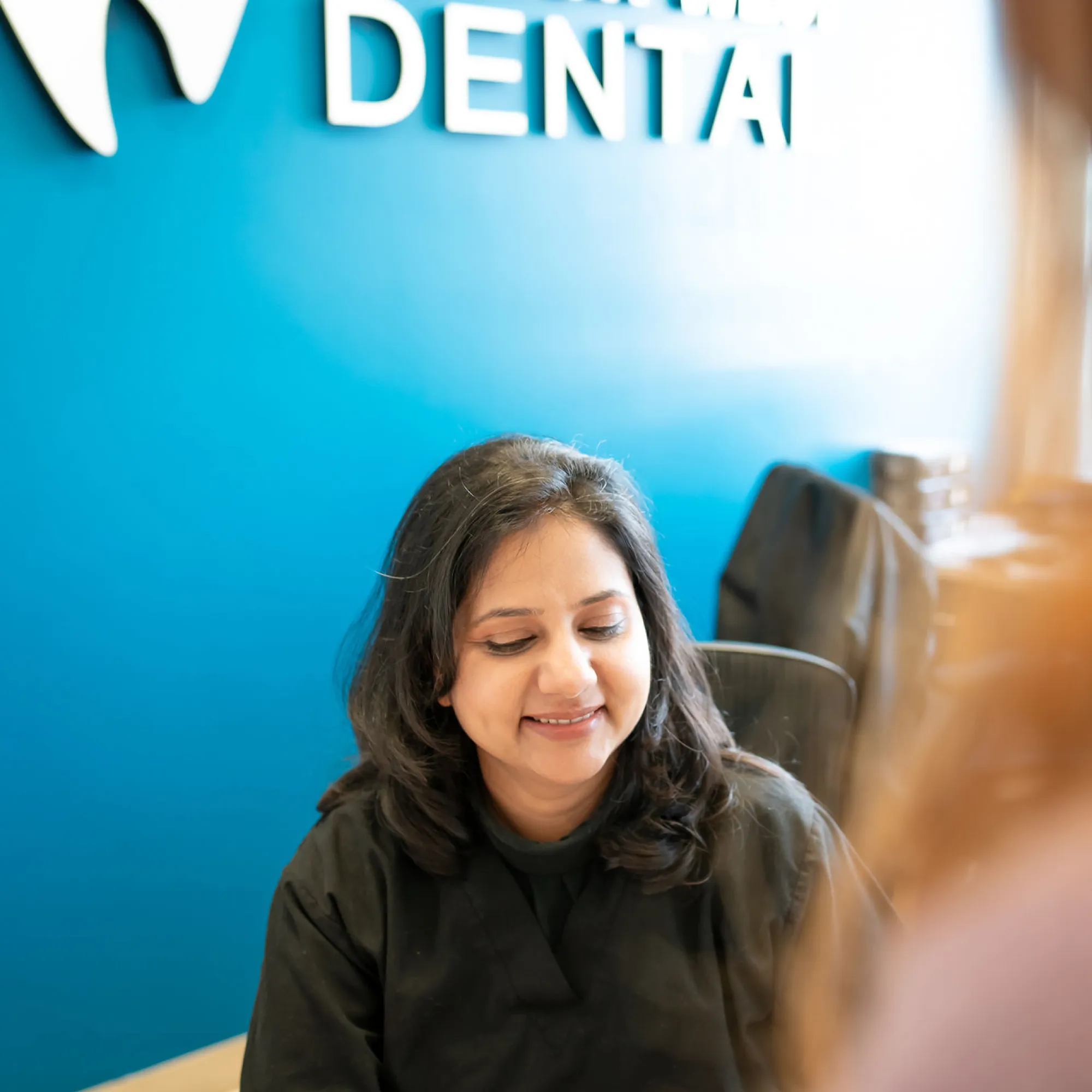 Smiling woman in black scrubs sitting at a desk against a blue wall with part of the word 'DENTAL' visible.