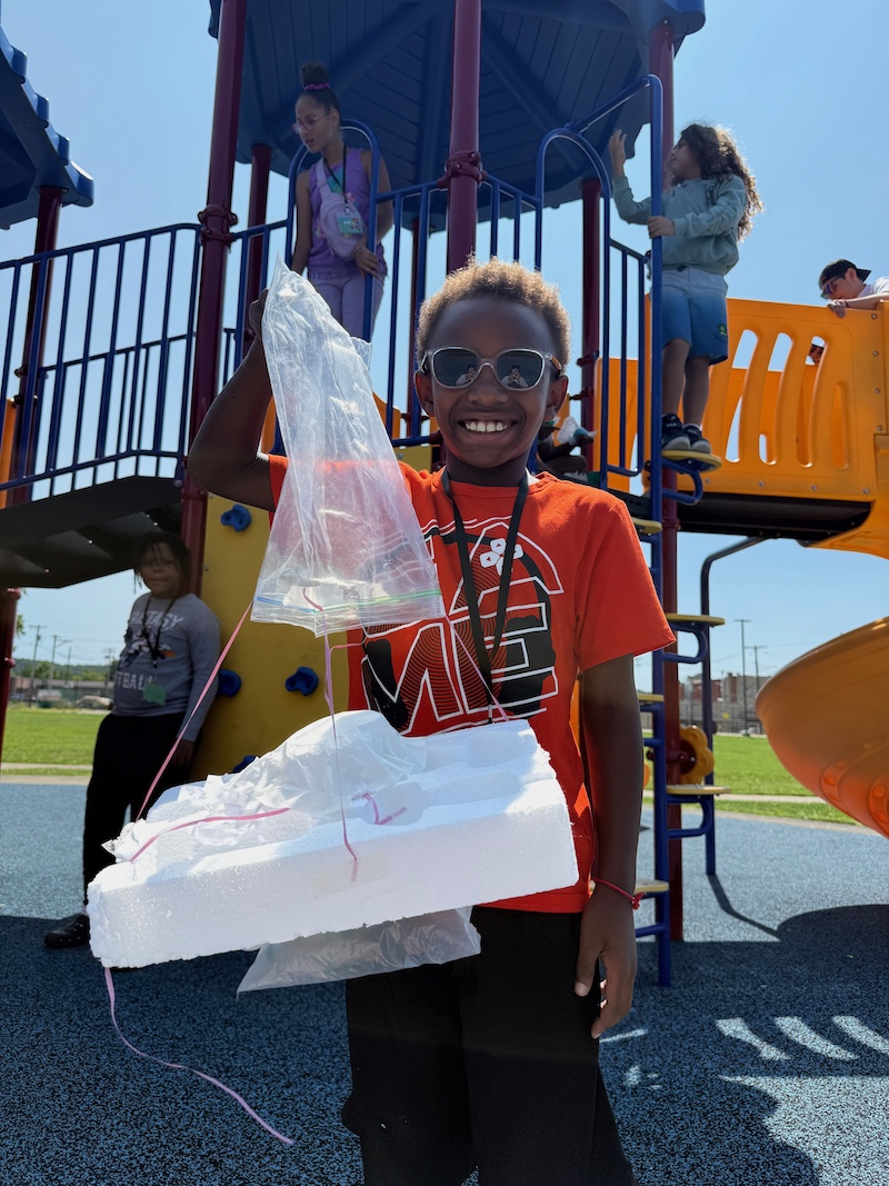 Student wearing an orange shirt and sunglasses stands in front of a playground.