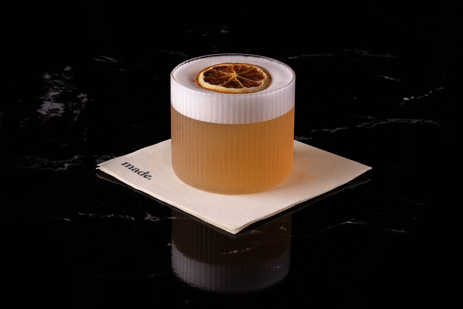 A crafted cocktail with a dried citrus garnish, served in a ribbed glass on a napkin embossed with the word “made.” against a black marble surface.