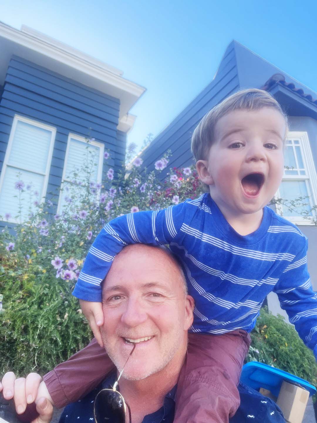 Smiling man carrying a joyful young boy wearing a blue striped shirt on his shoulders outside a house with purple flowers in the background.