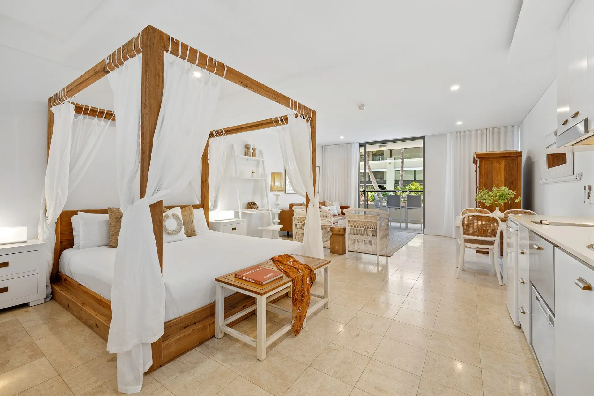 King-size four-poster bed with white muslin curtains in an open-plan studio apartment, with living area and balcony beyond