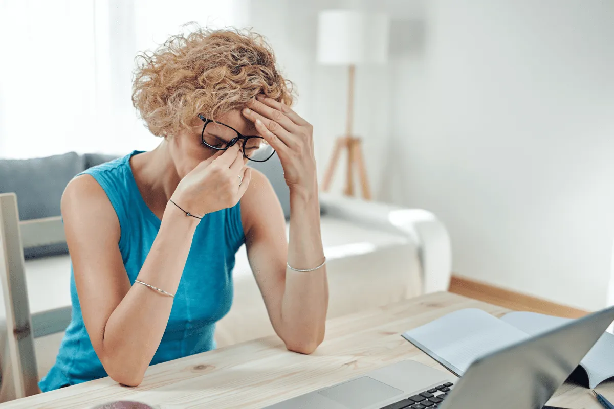 Person sitting at a desk with a laptop and notebook, holding their head in a stressful pose.