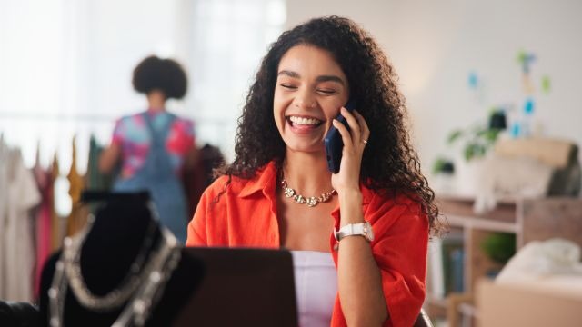 mulher de cabelos cacheados sorrindo ao telefone e olhando para um notebook em ambiente de loja