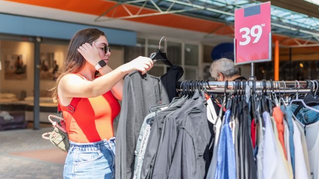mulher branca segurando um cabide com peça de roupa em frente a uma arara de roupas com placa indicando preço