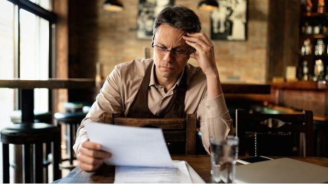 Homem branco de cabelos escuros, sentado à mesa de um bar, com a cabeça apoiada em uma das mãos enquanto analisa papéis