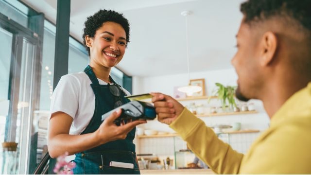 Mulher negra de cabelos curtos e cacheados, usando um avental, segura máquininha enquanto homem negro aproxima cartão