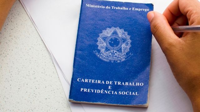 Carteira de Trabalho e Previdência Social azul sobre fundo claro, com mão segurando uma caneta ao lado