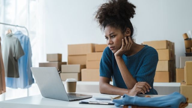 Mulher negra com cabelo preso, sentada à mesa com notebook aberto; com arara de roupas e caixas de papelão ao fundo
