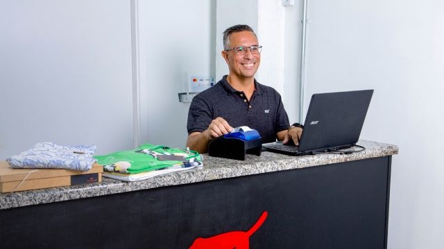 Homem de óculos sorrindo atrás de balcão utilizando notebook e impressora de etiquetas; sobre o balcão, roupas dobradas